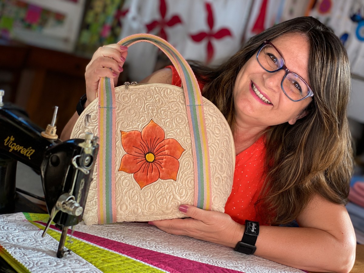 Márcia Baraldi sorrindo, segurando uma bolsa quiltada com uma flor laranja, ao lado da sua máquina de costura, num ateliê com quilts coloridos ao fundo